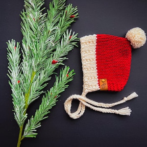 May include: A red and cream knitted baby bonnet with a cream pom-pom and ties. The bonnet features a small leather tag with the initials "WF". A sprig of evergreen with red berries is in the background.