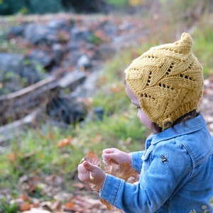 May include: A mustard yellow knitted hat with a pointed top and openwork pattern. The child is wearing a blue denim jacket. The background is a blurred outdoor scene with autumn leaves and greenery.