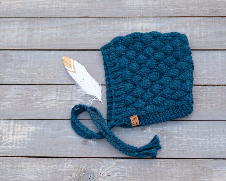 May include: Teal blue knitted hat with a textured pattern and a long tassel. The hat has a small brown leather tag with the letters "WF" on it.