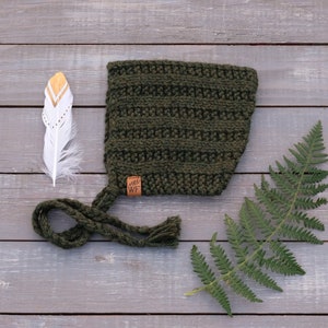 May include: A green crocheted baby bonnet with a brown leather tag that says "WF". The bonnet has a long tie that is tied in a knot. The bonnet is on a wooden surface with a white feather and a green fern.