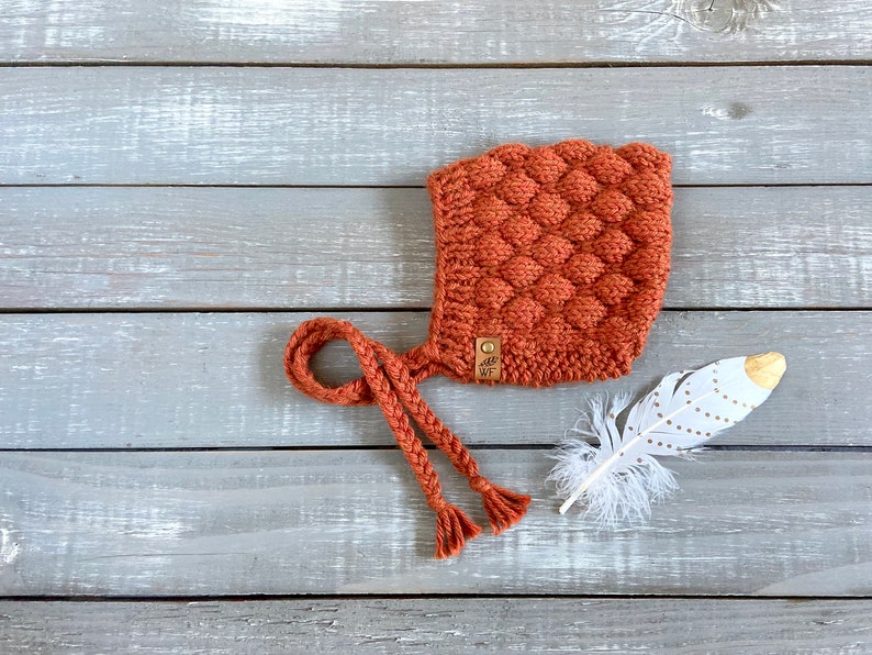 May include: A rust-colored knitted baby bonnet with a long tie and a small leather tag that says "WF". The bonnet is on a light gray wooden surface with a white feather with gold accents.