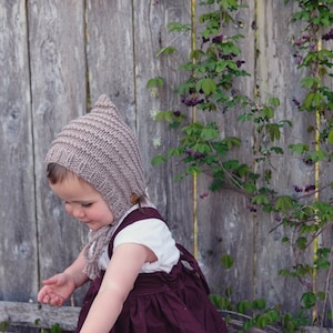 Peut inclure: Un enfant portant un bonnet en tricot marron avec un bouton en bois et une robe bordeaux, debout devant une clôture en bois avec des vignes vertes.