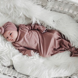 May include: A baby wearing a mauve knotted gown and a matching turban lies on a white fluffy blanket in a white wicker basket.