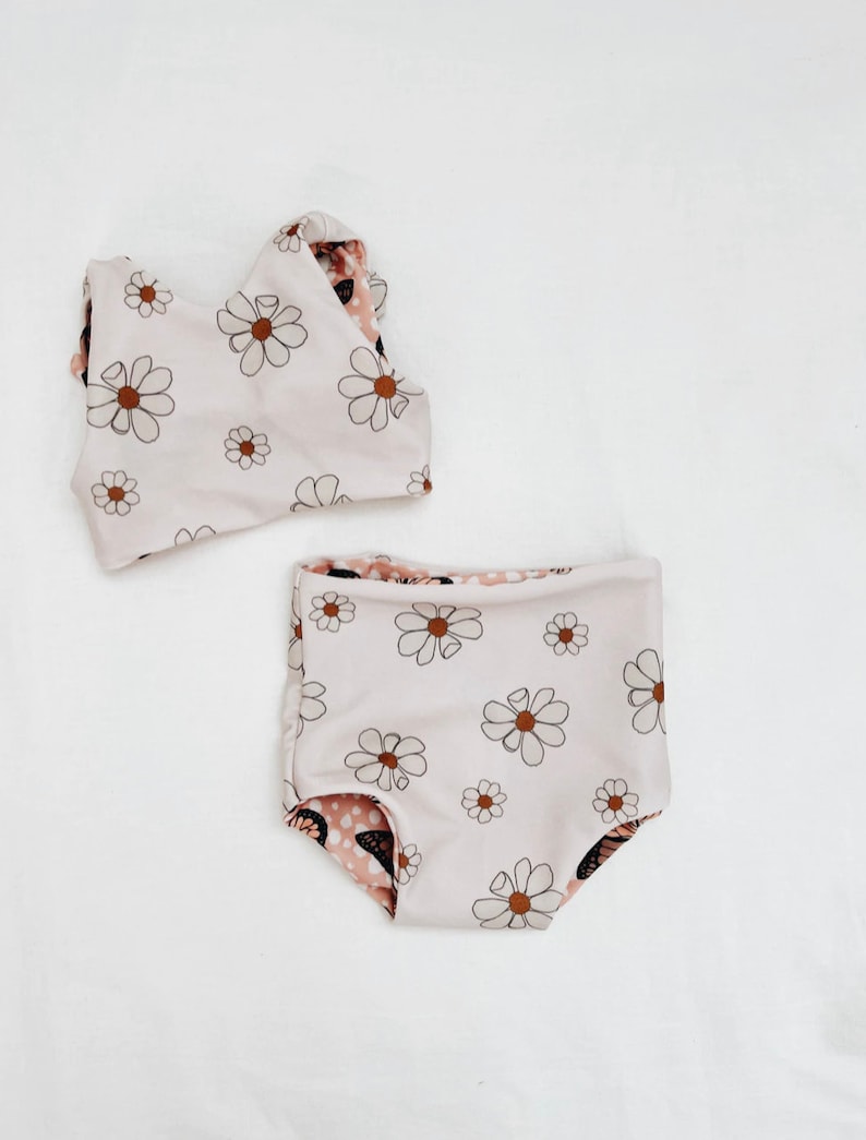 Peut inclure: Un maillot de bain deux pi&egrave;ces pour b&eacute;b&eacute;s et tout-petits. Le haut est un d&eacute;bardeur blanc &agrave; imprim&eacute; marguerite marron. Le bas est blanc avec un imprim&eacute; marguerite marron et un imprim&eacute; papillon marron &agrave; l'int&eacute;rieur.
