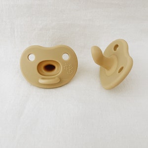 以下が含まれることがあります： ベージュのシリコン製ベビー用おしゃぶり2個。1つは平らに置かれ、もう1つは直立しています。平らに置かれたおしゃぶりには、側面に「#B」の文字があります。