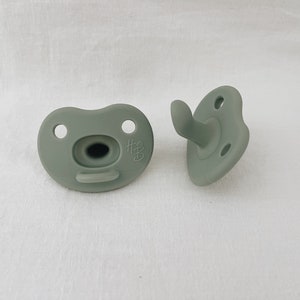 May include: Two sage green silicone pacifiers. One pacifier has a handle and the other has a small logo that says "#3".