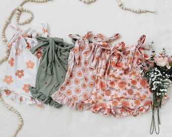 Puede incluir: Cuatro mamelucos con volantes para niña bebé con estampados florales. Los mamelucos son en tonos de blanco, verde y rosa con patrones florales naranjas. Los mamelucos tienen un lazo en los hombros.