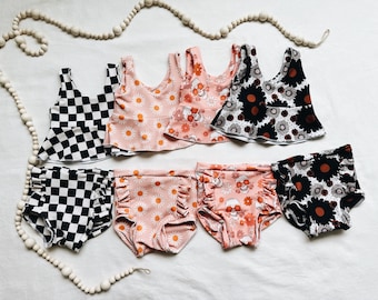 Peut inclure: Six paires de maillots de bain pour enfants avec différents motifs. Les motifs comprennent du carreau noir et blanc, du rose avec des marguerites blanches, du rose avec des crânes orange et du noir avec des marguerites blanches.