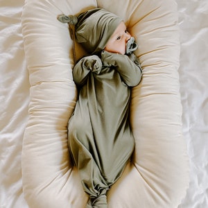 May include: A baby wearing a green knotted gown and matching hat lies in a cream-coloured nest. The baby is wearing a dummy.