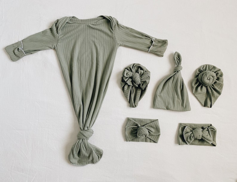 May include: Sage green baby gown and matching accessories set. The set includes a long-sleeved gown with a knotted bottom, two headbands, and three different style hats. The fabric has a ribbed texture and white trim.
