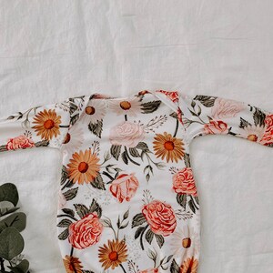 May include: A white baby gown with a floral print featuring pink roses, yellow daisies, and green leaves.