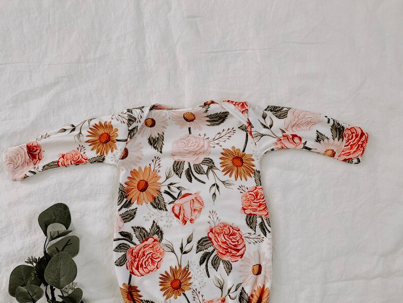 May include: A white baby gown with a floral print featuring pink roses, yellow daisies, and green leaves.