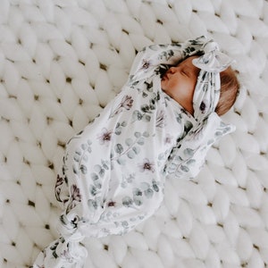 May include: A white swaddle with a floral pattern featuring eucalyptus leaves and small pink flowers. The swaddle is tied into a knot at the bottom and a matching headband is on the baby's head.