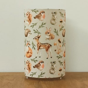Op de afbeelding: Een cilindrische lampenkap met een herhalend patroon van aquarel bosdieren, waaronder een hert, vos, konijn, egel, eekhoorn en roodborstje, afgewisseld met kleine paddenstoelen en groen gebladerte op een lichtcrème achtergrond.