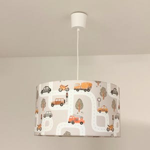 Op de afbeelding: Een cilindervormige hanglamp met een lichtgrijze lampenkap voorzien van een herhalend patroon van oranje en grijze voertuigen, waaronder brandweerwagens, politieauto's en motorfietsen, samen met witte kronkelende wegen en bomen.