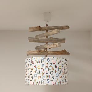Op de afbeelding: Een plafondlamp met een cilindrische lampenkap met een witte achtergrond bedrukt met een kleurrijk alfabetpatroon. Boven de kap zijn vijf lichtbruine stukken drijfhout horizontaal gestapeld en opgehangen aan een centraal koord.