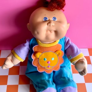 May include: A doll with a round face, dimpled cheeks, and blue eyes. It has curly red hair tied with a yellow flower accessory. The doll wears a blue long-sleeved outfit with purple sleeves, yellow trim, and a large orange lion graphic on the chest.
