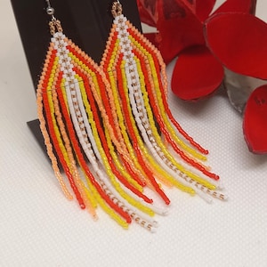 Puede incluir: Un par de aretes largos de cuentas con un diseño de flecos. Los aretes están hechos con cuentas blancas, naranjas, amarillas y rojas.