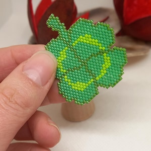 May include: A beaded shamrock in shades of green and yellow. The four-leaf clover is made of small, square beads. The beaded shamrock is held in a person's hand. The background is blurred, with hints of red and brown.
