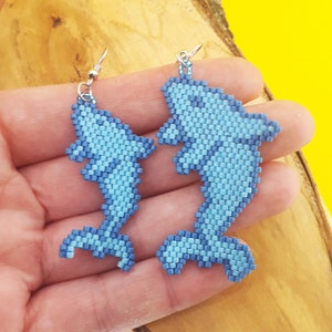 Könnte beinhalten: Ein Paar blaue Perlen-Delfin-Ohrringe. Die Ohrringe bestehen aus kleinen blauen Perlen und sind in Form von Delfinen geformt. Die Ohrringe hängen an silbernen Haken.