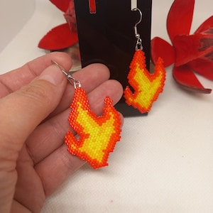 May include: A pair of flame-shaped earrings crafted from small, vibrant beads in shades of bright yellow, orange, and red. Each earring features a silver-toned metal hook for attachment, with the beaded flame design hanging vertically from the wire.