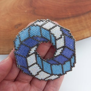 May include: A blue, white, and purple beaded circle with a hole in the center. The circle is made of small, square beads that are sewn together.