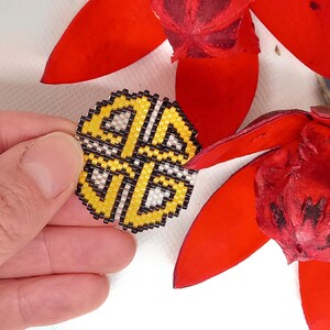 May include: A yellow, black, and white beaded round ornament with a geometric design. The ornament is being held by a hand.