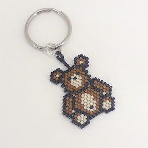 May include: A brown and white beaded bear keychain with black accents. The bear is attached to a silver key ring.