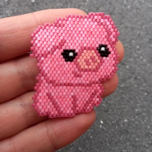 Pode incluir: Um porco de contas cor-de-rosa com olhos pretos e um focinho rosa. O porco é feito de pequenas contas quadradas e tem cerca de 5 cm de altura.