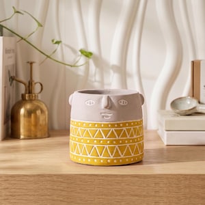 May include: A decorative ceramic planter with a face design. The top is light gray, and the bottom is yellow with a white geometric pattern. The planter has small handles and is suitable for small plants. Other items include books and a brass mister.