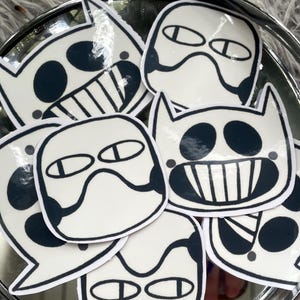 May include: A selection of white stickers with black outlines and details, depicting cartoon faces with diverse expressions. The stickers are arranged on a silver, circular tray, creating a playful and artistic display.
