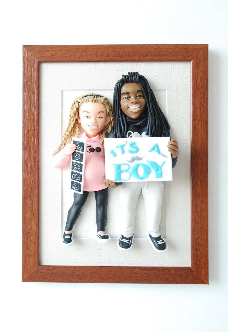 May include: A framed 3D figurine of a couple holding an ultrasound image and a sign that says "It's a Boy!" The figurine is made of clay and is painted in realistic colors. The frame is made of wood and has a brown finish.