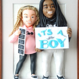 May include: A clay figurine of a couple holding a sign that says "It's a Boy!" The woman is holding an ultrasound picture and the man has a mustache on his sign.