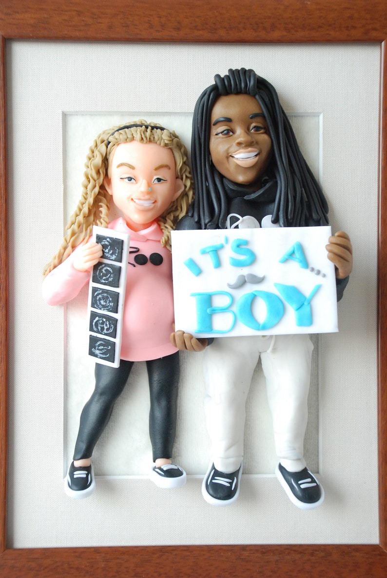 May include: A clay figurine of a couple holding a sign that says "It's a Boy!" The woman is holding an ultrasound picture and the man has a mustache on his sign.
