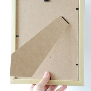 May include: A light wood picture frame with a cardboard backing. The frame is being held by a hand.
