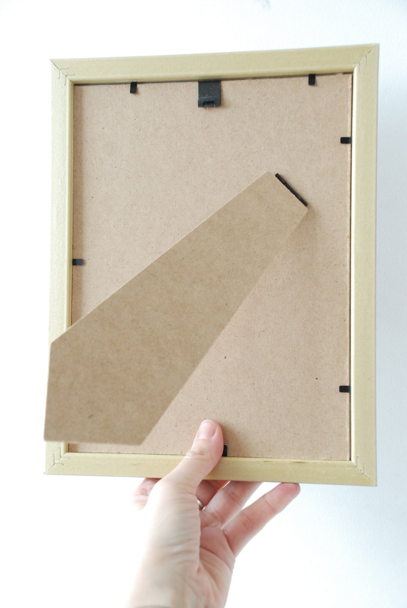 May include: A light wood picture frame with a cardboard backing. The frame is being held by a hand.