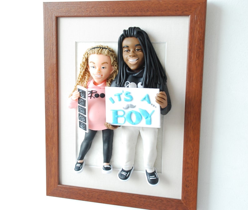 May include: A framed 3D clay sculpture of a couple holding a sign that says "It's a Boy!" The woman is holding an ultrasound picture and the man is wearing a black hoodie.