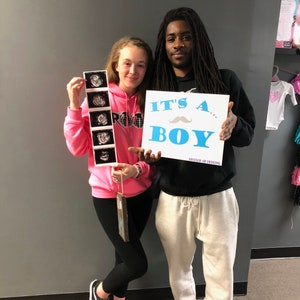 May include: A couple holding up a sign that says "It's a... Boy" with a blue mustache. The woman is holding a strip of ultrasound images. The man is wearing a black hoodie and white sweatpants. The woman is wearing a pink hoodie and black leggings.