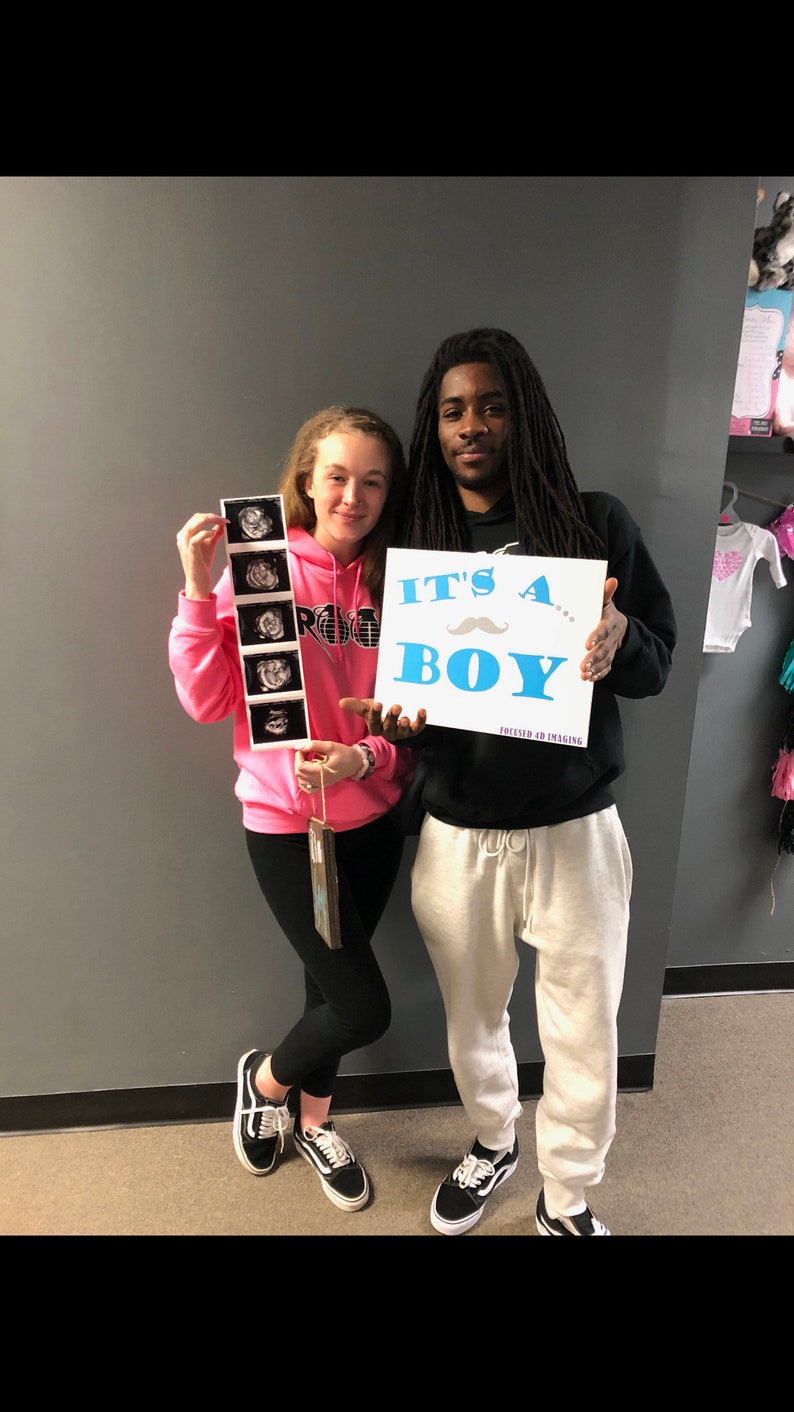 May include: A couple holding up a sign that says "It's a... Boy" with a blue mustache. The woman is holding a strip of ultrasound images. The man is wearing a black hoodie and white sweatpants. The woman is wearing a pink hoodie and black leggings.