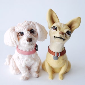 Może przedstawiać: Dwie figurki psów, biały pudel z różową obrożą i beżowa chihuahua z brązową obrożą, siedzą obok siebie. Oba psy mają szczegółowe rysy twarzy i wyraziste oczy, na białym tle.