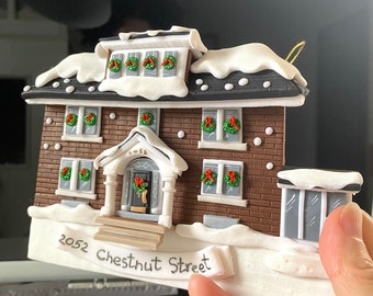 May include: A brown brick house with white snow on the roof and a white sign that reads "2052 Chestnut Street". The house has a red door with a wreath and a white porch with steps. The house is decorated with green wreaths and red bows.