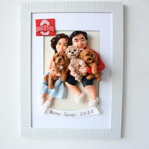 Könnte beinhalten: Eine gerahmte Tonskulptur eines Paares mit drei Hunden. Das Paar hält zwei der Hunde, und der dritte Hund sitzt auf dem Boden. Der Rahmen ist weiß mit einem weißen Passepartout. Das Paar trägt Freizeitkleidung. Die Hunde sind braun und weiß. Der Rahmen hat einen rot-weißen Ohio State-Patch in der oberen Ecke. Der Rahmen hat auch ein weißes Banner mit dem Text "Moore-Gongs 2022".