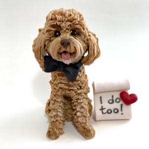 May include: A detailed figurine of a light brown dog with a black bow tie, tongue out, and a small sign that says "I do too!" with a red heart. The dog is sitting down.