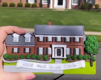 May include: A miniature house ornament with a brick facade, gray roof, and white trim. The ornament has a white banner with the text "6008 West Grove Circle" written in black marker.
