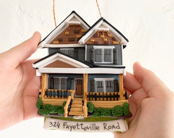 May include: A handmade clay ornament of a two-story house with a brown brick facade, white trim, and a gray front door. The house has a porch with a wooden railing and steps leading up to the front door. The ornament is decorated with green bushes and a sign that reads "324 Fayetteville Road".