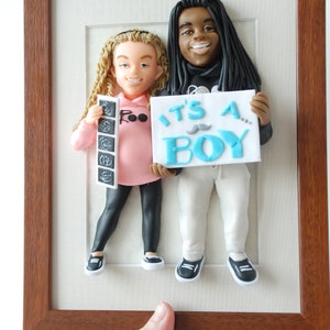 May include: A framed 3D clay sculpture of a couple holding an "It's a Boy" sign. The woman is holding a stack of ultrasound images. The sculpture is set against a white background.