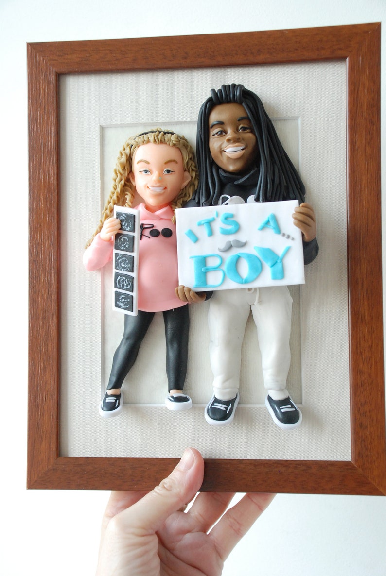 May include: A framed 3D clay sculpture of a couple holding an "It's a Boy" sign. The woman is holding a stack of ultrasound images. The sculpture is set against a white background.