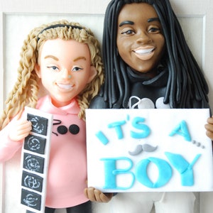 May include: A clay figurine of a couple holding a sign that says "It's a Boy!" The woman is holding a stack of ultrasound images. The man has dreadlocks and is wearing a black hoodie.