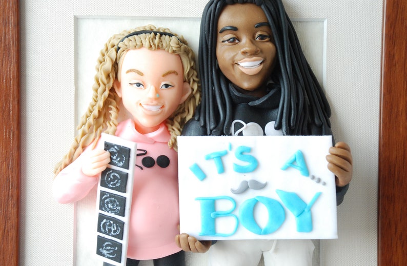 May include: A clay figurine of a couple holding a sign that says "It's a Boy!" The woman is holding a stack of ultrasound images. The man has dreadlocks and is wearing a black hoodie.