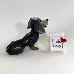 Pode incluir: Uma figurinha de dachshund preta e branca com o nariz castanho e a língua de fora, olhando para um rolo branco com o texto "I do too!" e um coração vermelho.